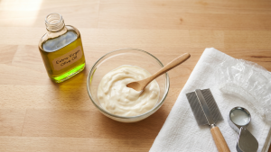 An editorial flat-lay on a light wood kitchen counter showing a glass bowl of full-fat mayonnaise with a wooden spoon, a small bottle of extra-virgin olive oil, a folded white cotton towel with a fine-tooth metal nit comb, a clear shower cap, and a handheld magnifier, under soft morning daylight.