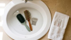 A clean white bathroom sink with two family hairbrushes and one fine-tooth metal nit comb submerged in clear hot water, a folded white cotton towel and a small thermometer on the counter beside it, under soft natural daylight.