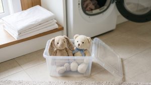 A clear plastic storage bin holding two soft pastel stuffed animals (a bunny and a teddy bear) on a home laundry-room floor, with a folded white bed sheet and a dryer drum softly out of focus in the background, under calm natural daylight.