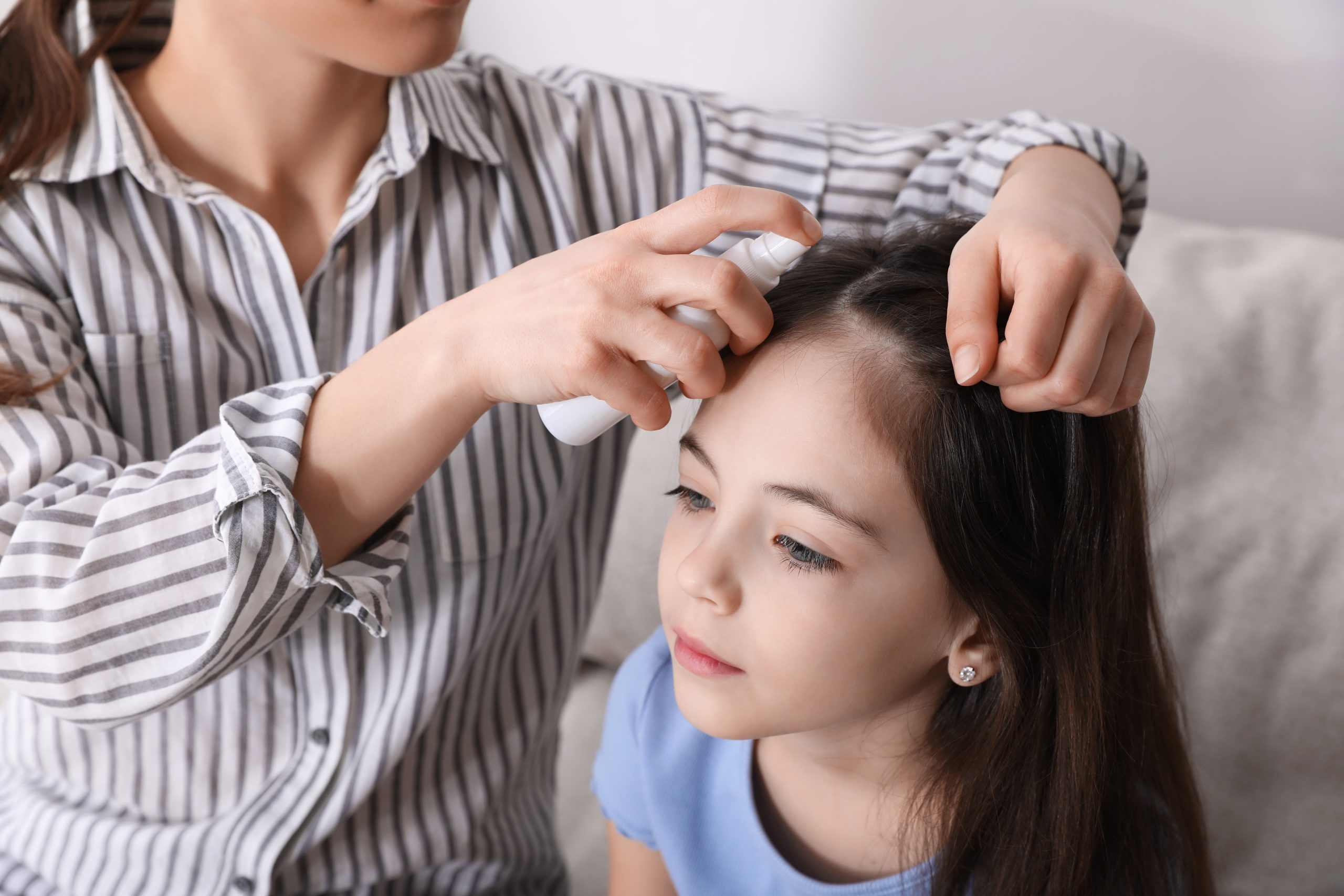 Mother applying a leave-in prevention spray to her daughter's hair indoors before school during a classroom lice outbreak window.
