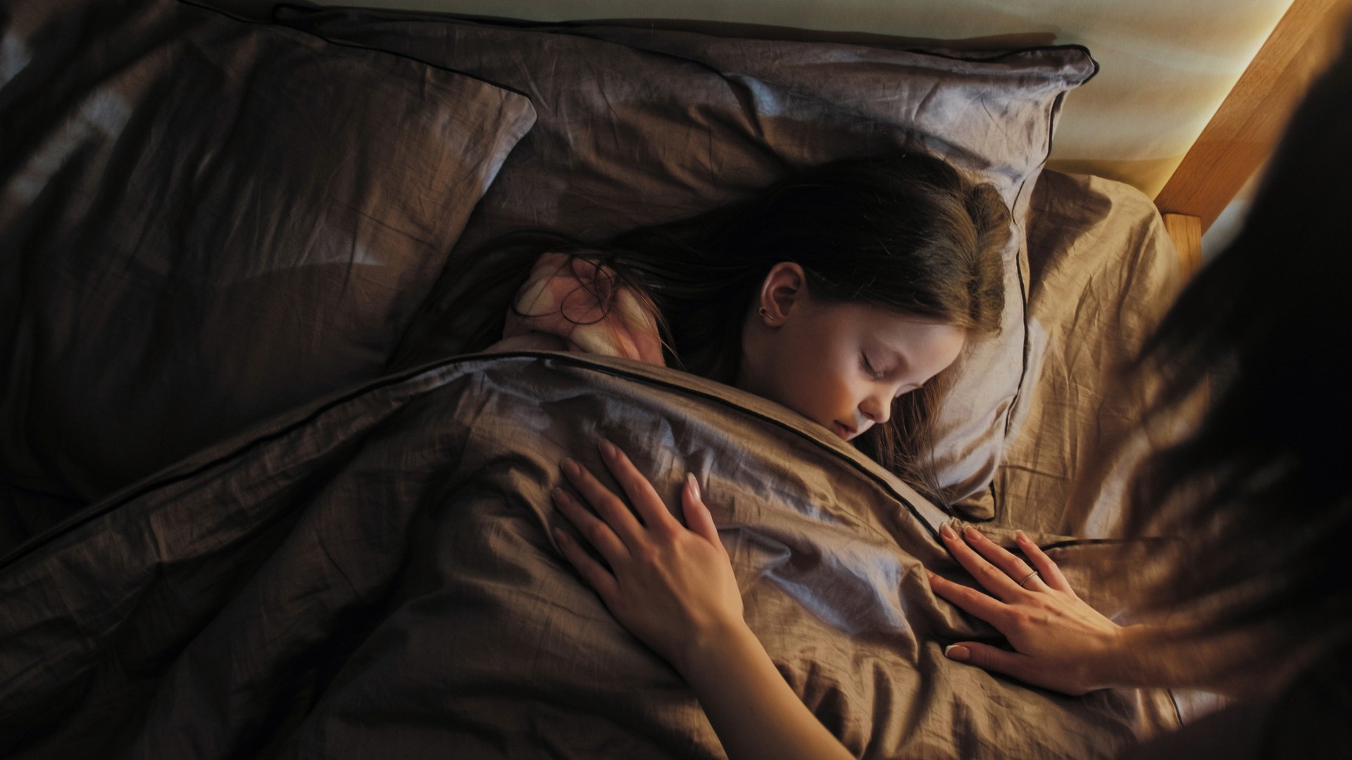 A mother gently covers her sleeping daughter with a blanket in a calm dark bedroom at night.