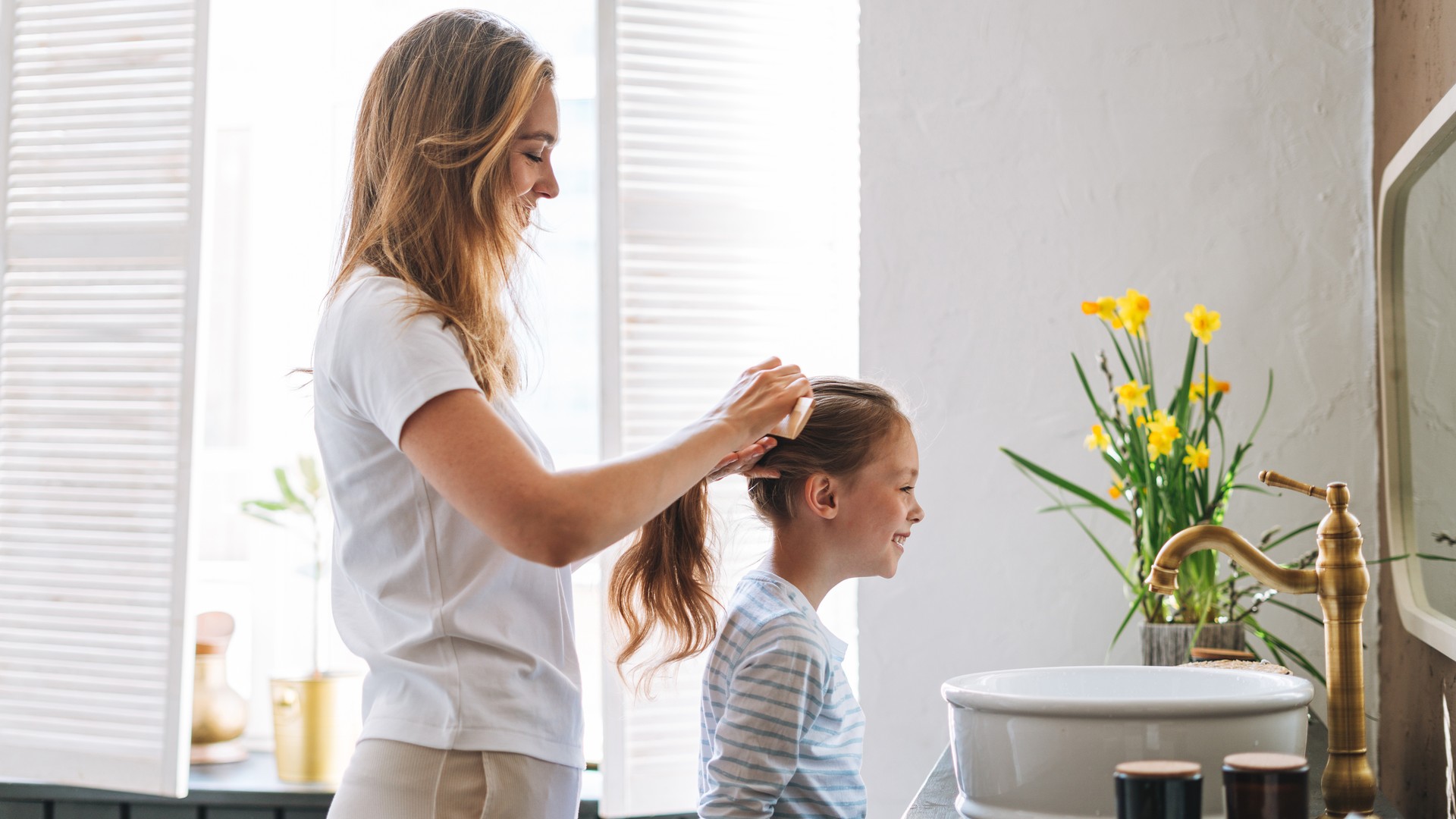 A parent at a bright home bathroom sink running a fine-tooth metal nit comb slowly through a section of a child's damp, conditioned hair, wiping the comb on a white paper towel between passes during an at-home head lice check