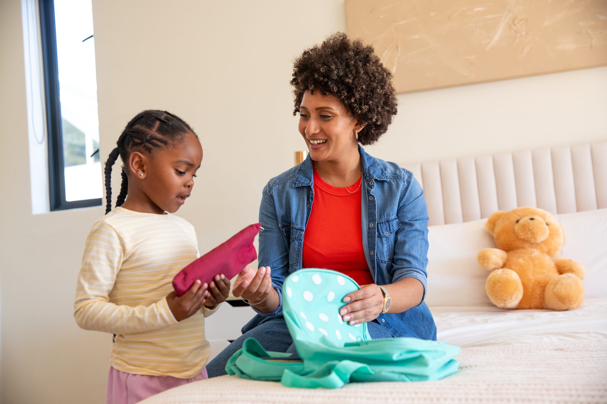 A calm parent and school-aged daughter packing a blue polka-dot duffel-style backpack on a neatly made bed in a sunlit bedroom next to a teddy bear, representing the calm parent response between receiving a summer camp lice notification and pickup day