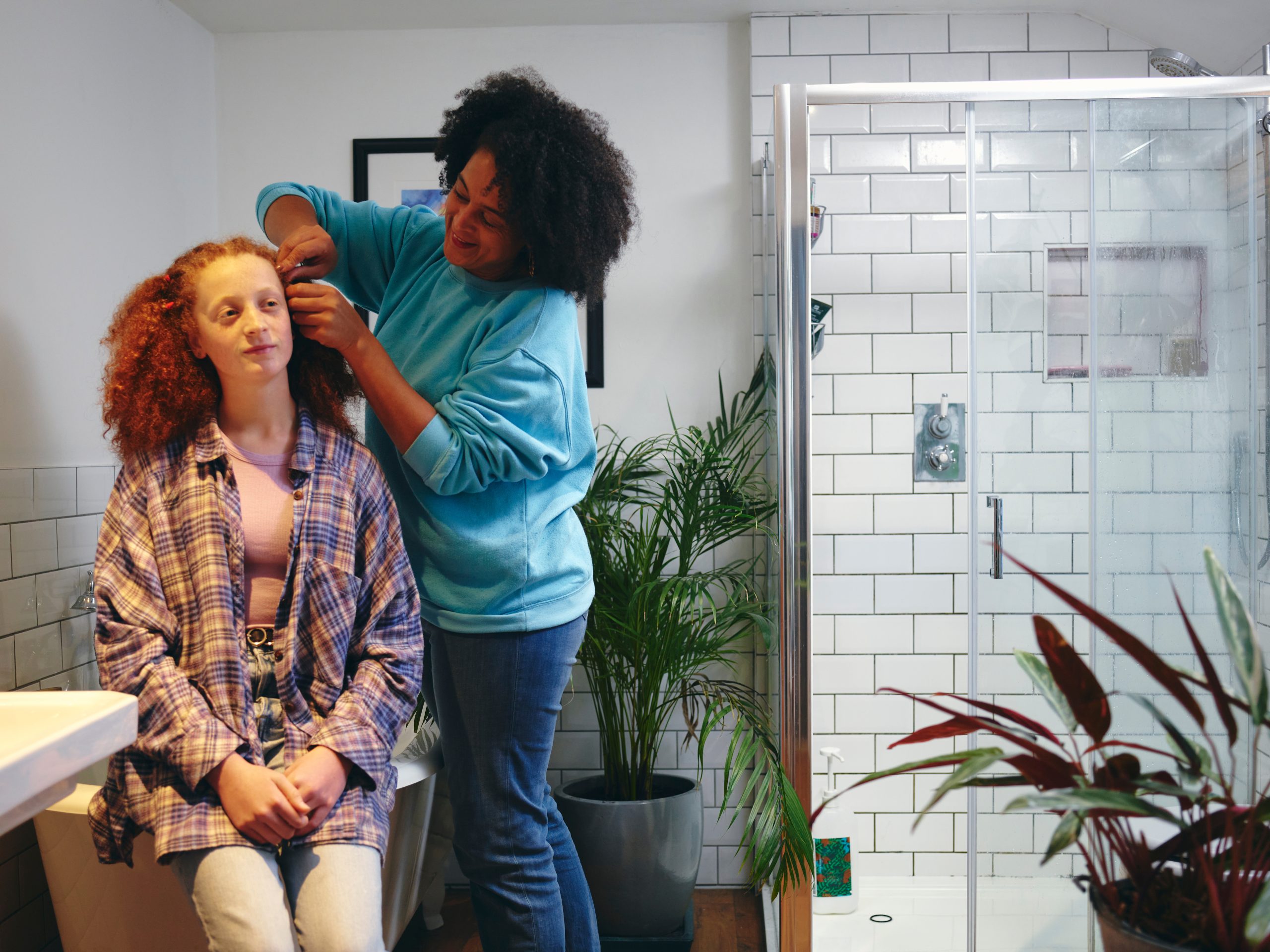 A person arranges the curly red hair of someone seated in a bathroom.
