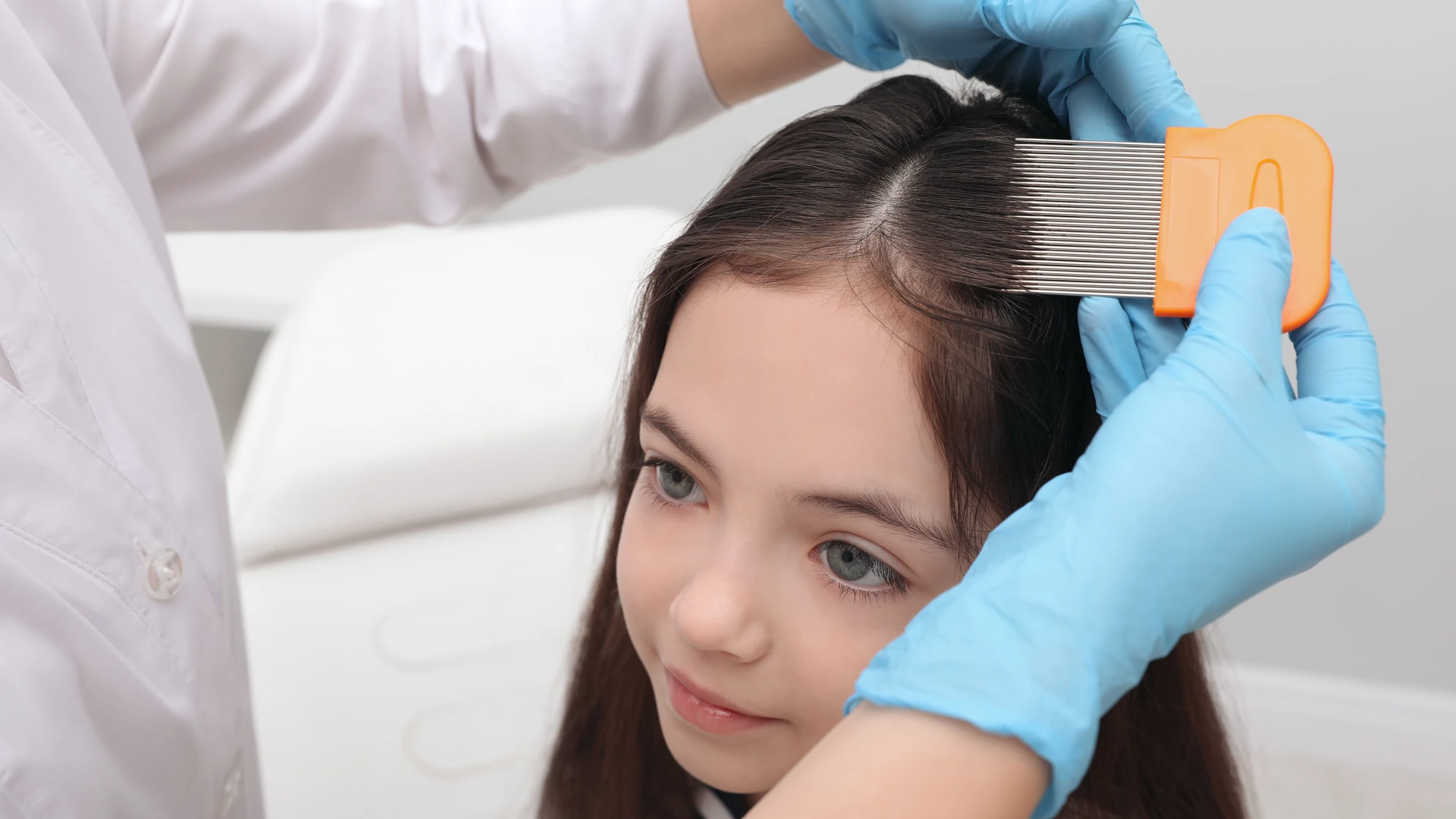 Blue-gloved hands hold an orange-handled fine-toothed comb in dark hair beside the part.