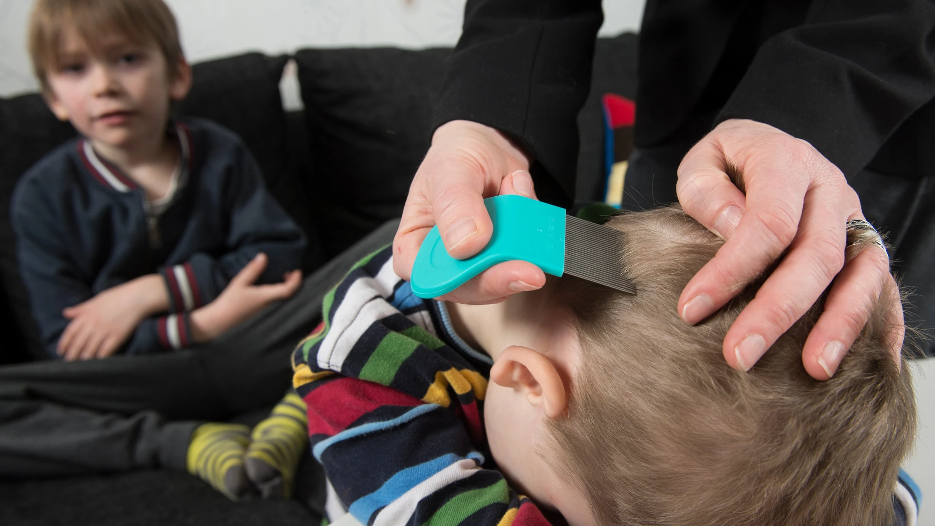 Hands comb short light hair with a turquoise-handled fine-toothed comb while another person sits nearby.